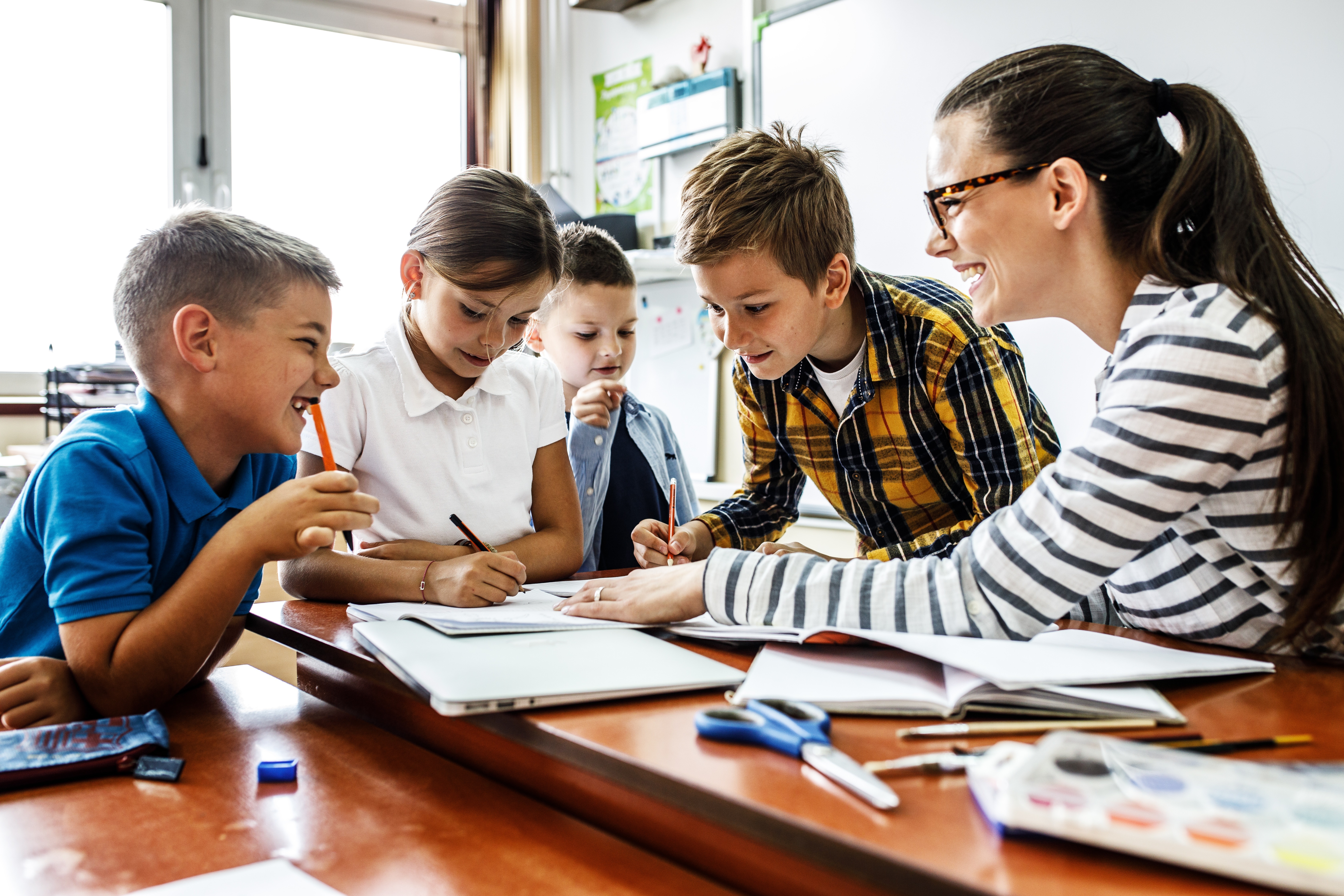 Female teacher helps school kids to finish they lesson.They sitting all together at one desk.